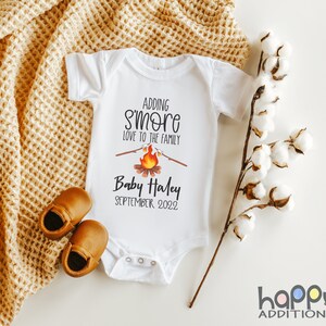 May include: White baby onesie with the text "Adding s'more love to the family" and a campfire graphic. The onesie also has the text "Baby Haley September 2022".
