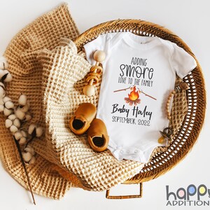 May include: White onesie with the text "Adding s'more love to the family" and a campfire graphic. Below the text is "Baby Haley September 2022". The onesie is in a wicker basket with a brown blanket, wooden toys, and brown baby shoes.