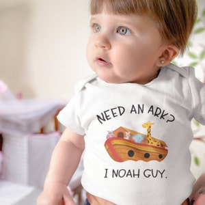 May include: A white baby onesie with a cartoon illustration of Noah's Ark with animals on board. The text on the onesie reads "Need an ark? I Noah guy."