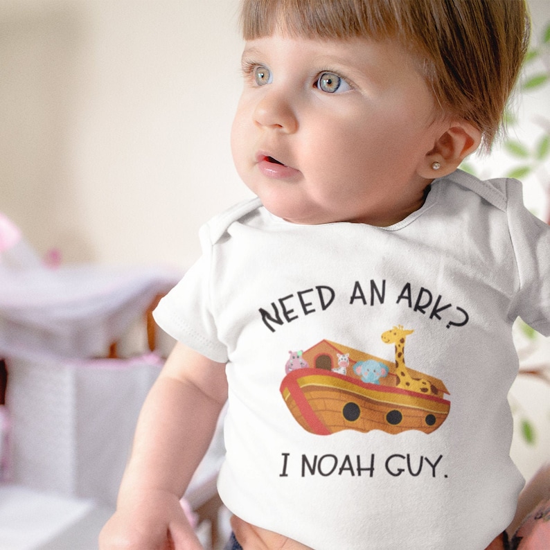 May include: A white baby onesie with a cartoon illustration of Noah's Ark with animals on board. The text on the onesie reads "Need an ark? I Noah guy."