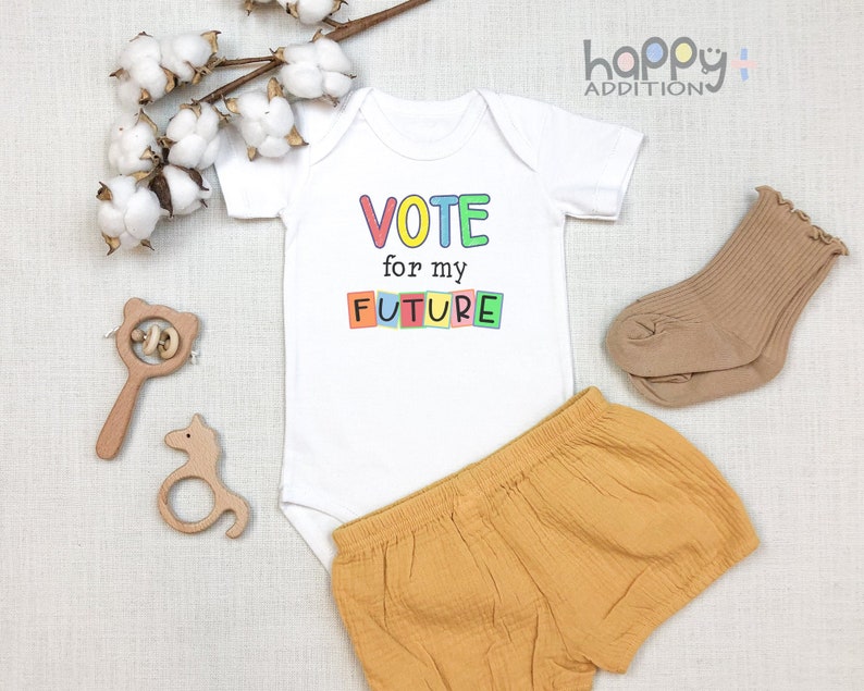 May include: White baby onesie with the text "VOTE for my FUTURE" in colorful letters. The onesie is paired with mustard yellow baby shorts and brown socks.