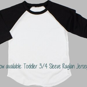 Pu&ograve; includere: Maglietta bianca da bambino a 3/4 di manica con maniche nere. La maglietta ha un collo rotondo e una vestibilit&agrave; rilassata. "Now available: Toddler 3/4 Sleeve Raglan Jersey!" &egrave; scritto nella parte inferiore dell'immagine.