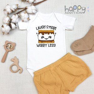 Pu&ograve; includere: Body bianco per neonato con maniche corte e il testo "Laugh S'more Worry Less!" in nero. Un s'more di cartone animato con un viso sorridente &egrave; sopra il testo. Il body &egrave; abbinato a pantaloncini marroni.