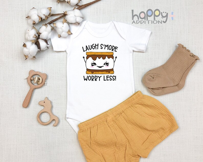 Pu&ograve; includere: Body bianco per neonato con maniche corte e il testo "Laugh S'more Worry Less!" in nero. Un s'more di cartone animato con un viso sorridente &egrave; sopra il testo. Il body &egrave; abbinato a pantaloncini marroni.