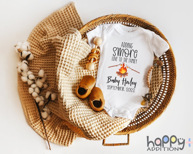 May include: White onesie with the text "Adding s'more love to the family" and a campfire graphic. Below the text is "Baby Haley September 2022". The onesie is in a wicker basket with a brown blanket, wooden toys, and brown baby shoes.