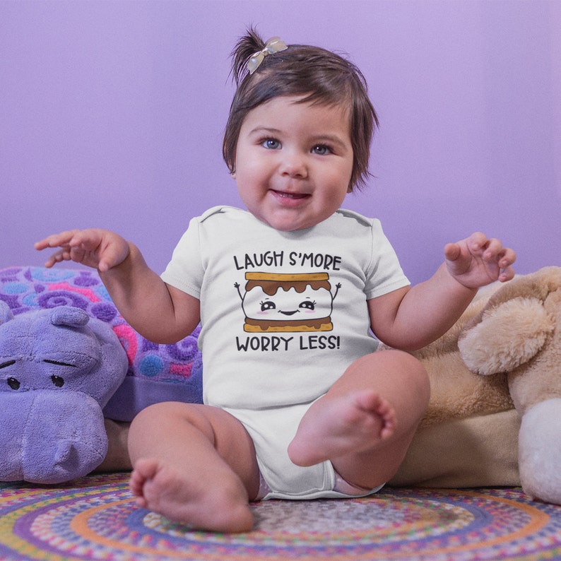 Pu&ograve; includere: Un body bianco per neonato con un grafico di s'more a cartone animato e il testo "Laugh S'more Worry Less!" Un bambino indossa il body e siede su un tappeto colorato.