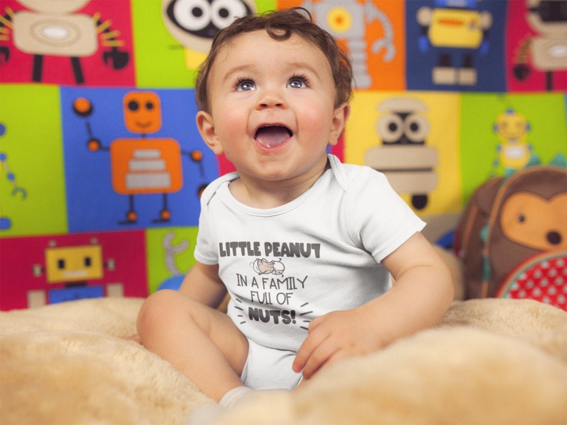 May include: A white baby onesie with the text "Little Peanut in a family full of nuts!" and a cartoon peanut in a shell. A baby is wearing the onesie and sitting on a light brown surface.