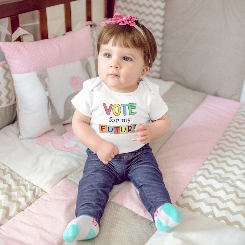 May include: A baby wearing a white onesie with the text "VOTE for my FUTURE" in colorful block letters. The baby is sitting on a pink and gray patterned crib mattress.
