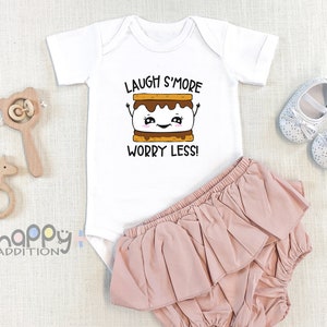 Pu&ograve; includere: Body bianco per neonato con un grafico di marshmallow arrostito a cartone animato e il testo "Laugh S'more Worry Less!" Il body &egrave; abbinato a un pannolino con volant rosa.