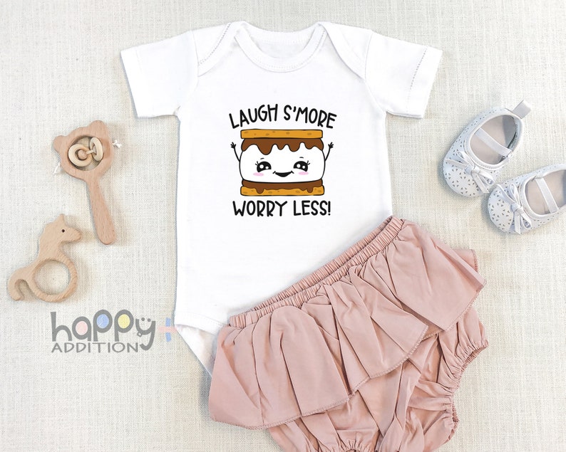Pu&ograve; includere: Body bianco per neonato con un grafico di marshmallow arrostito a cartone animato e il testo "Laugh S'more Worry Less!" Il body &egrave; abbinato a un pannolino con volant rosa.