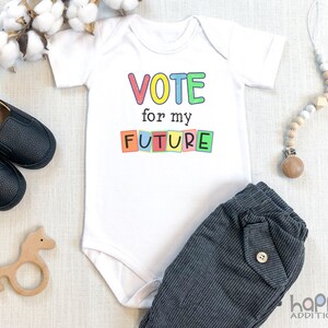 May include: White baby onesie with the text "VOTE for my FUTURE" in colorful block letters. The onesie is on a white surface with a wooden toy horse, a pair of black baby shoes, and a wooden bead necklace.