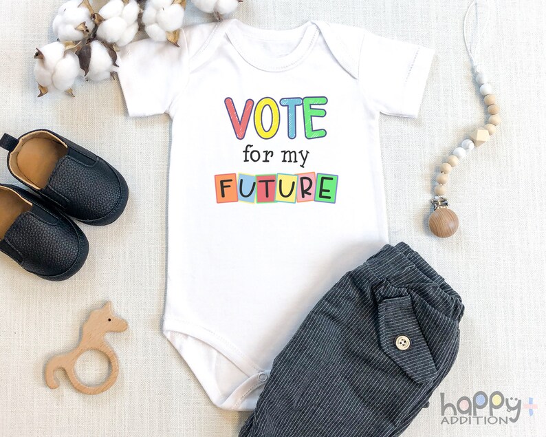 May include: White baby onesie with the text "VOTE for my FUTURE" in colorful block letters. The onesie is on a white surface with a wooden toy horse, a pair of black baby shoes, and a wooden bead necklace.