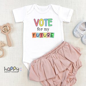 May include: White baby onesie with the text "VOTE for my FUTURE" in colorful block letters. The onesie is paired with a pink ruffled diaper cover.