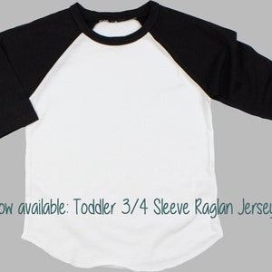 May include: White toddler 3/4 sleeve raglan jersey with black sleeves. The jersey has a rounded neckline and a relaxed fit. "Now available: Toddler 3/4 Sleeve Raglan Jersey!" is written in the bottom of the image.