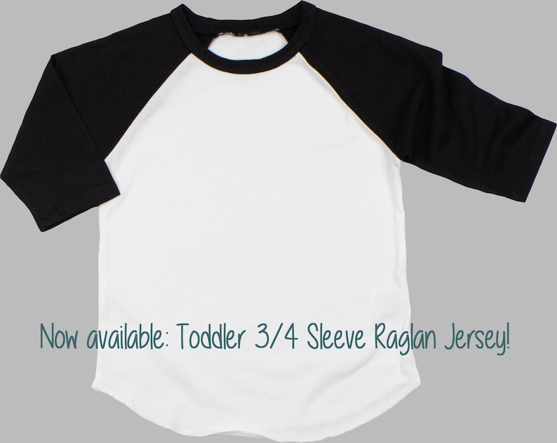 May include: White toddler 3/4 sleeve raglan jersey with black sleeves. The jersey has a rounded neckline and a relaxed fit. "Now available: Toddler 3/4 Sleeve Raglan Jersey!" is written in the bottom of the image.