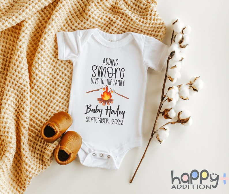 May include: White baby onesie with the text "Adding s'more love to the family" and a campfire graphic. The onesie also has the text "Baby Haley September 2022".