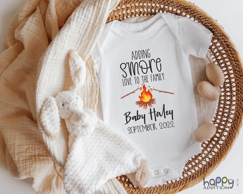 May include: White baby onesie with a campfire graphic and the text "Adding s'more love to the family" and "Baby Haley September 2022".