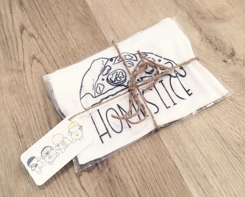May include: A white t-shirt with a black and white illustration of a pizza slice and the text "Home Slice". The t-shirt is wrapped in brown paper and tied with twine. A small tag with a cartoon illustration of four people is attached to the package.