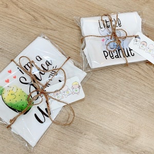 May include: Two white baby onesies wrapped in brown twine and clear plastic. One onesie has a cactus and the text "I'm a little cactus". The other onesie has an elephant and the text "little peanut".