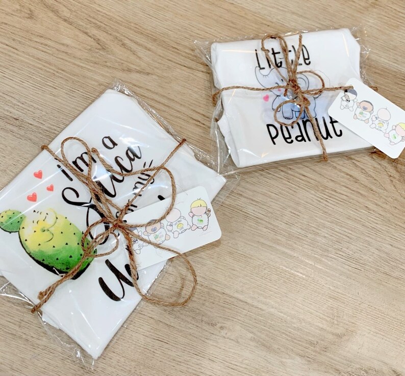 May include: Two white baby onesies wrapped in brown twine and clear plastic. One onesie has a cactus and the text "I'm a little cactus". The other onesie has an elephant and the text "little peanut".