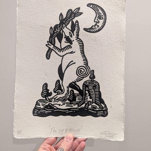 May include: A black and white linocut print depicting a rabbit-like figure holding a leafy branch, with a crescent moon and a tree stump. The text "The Pig of Wands" is below the image. The artwork is on textured paper.
