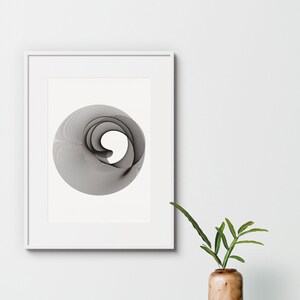 Op de afbeelding: Een zwart-wit abstract geometrisch ontwerp in een wit frame op een witte muur. Een houten vaas met een groene plant staat op de voorgrond.