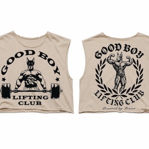 May include: A beige sleeveless crop top featuring a black graphic print. The front displays a muscular dog lifting a barbell with the text GOOD BOY LIFTING CLUB. The back shows a muscular dog flexing inside a laurel wreath with the same text.