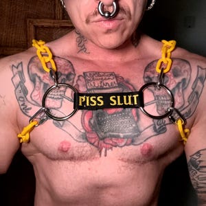 May include: A chest harness featuring bright yellow plastic chain links connected to two large silver-toned metal rings. A central black fabric strap is embroidered with the yellow text PISS SLUT, positioned horizontally between the two metal rings.
