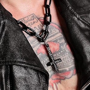 Peut inclure: Un collier à chaîne noire avec un pendentif en forme de croix. Le pendentif porte le mot "DEVIANT" et d'autres textes. Le collier est porté sur une veste en cuir noire.