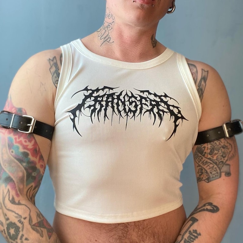 Op de afbeelding: Een wit geribbeld tanktop met een zwarte grafische afbeelding met de tekst "F.A.U.S.T." Het tanktop is gecropped en heeft een racerback-stijl.