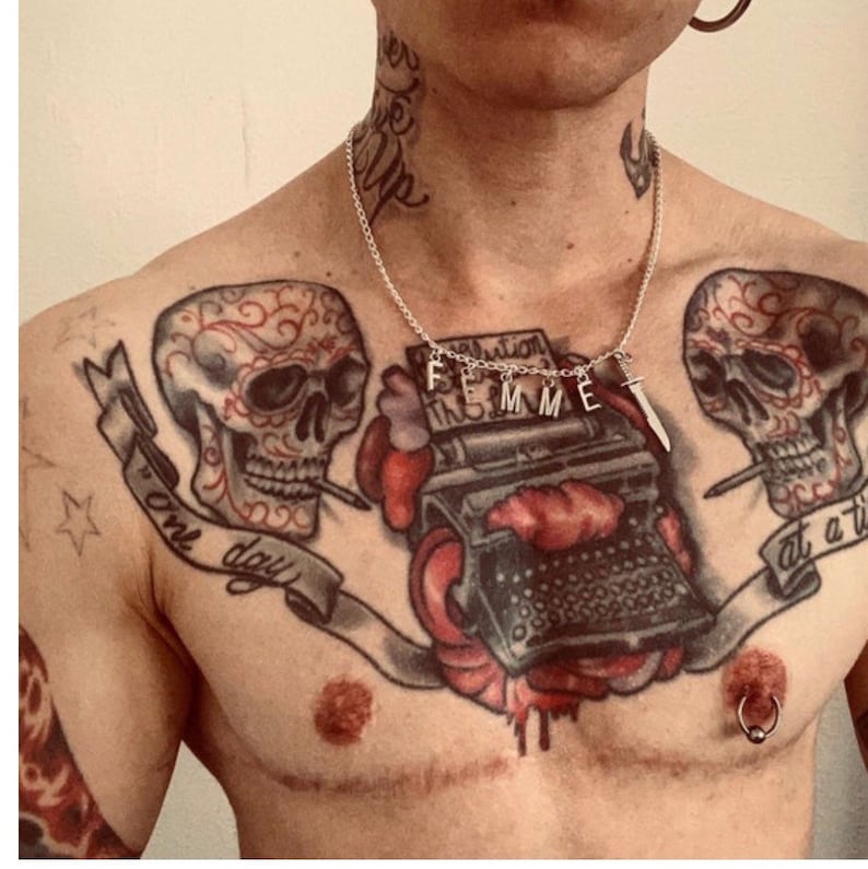 May include: A person with several tattoos on their chest, including two sugar skulls, a typewriter, and a banner that reads "one day at a time". They are wearing a silver necklace with the word "FEMME" spelled out in charms and a small dagger charm.