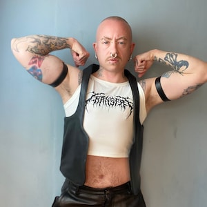Op de afbeelding: Een persoon draagt een zwart leren vest over een wit crop top met een zwarte grafische print. Ze dragen een zwarte leren broek en hebben tatoeages op hun armen.