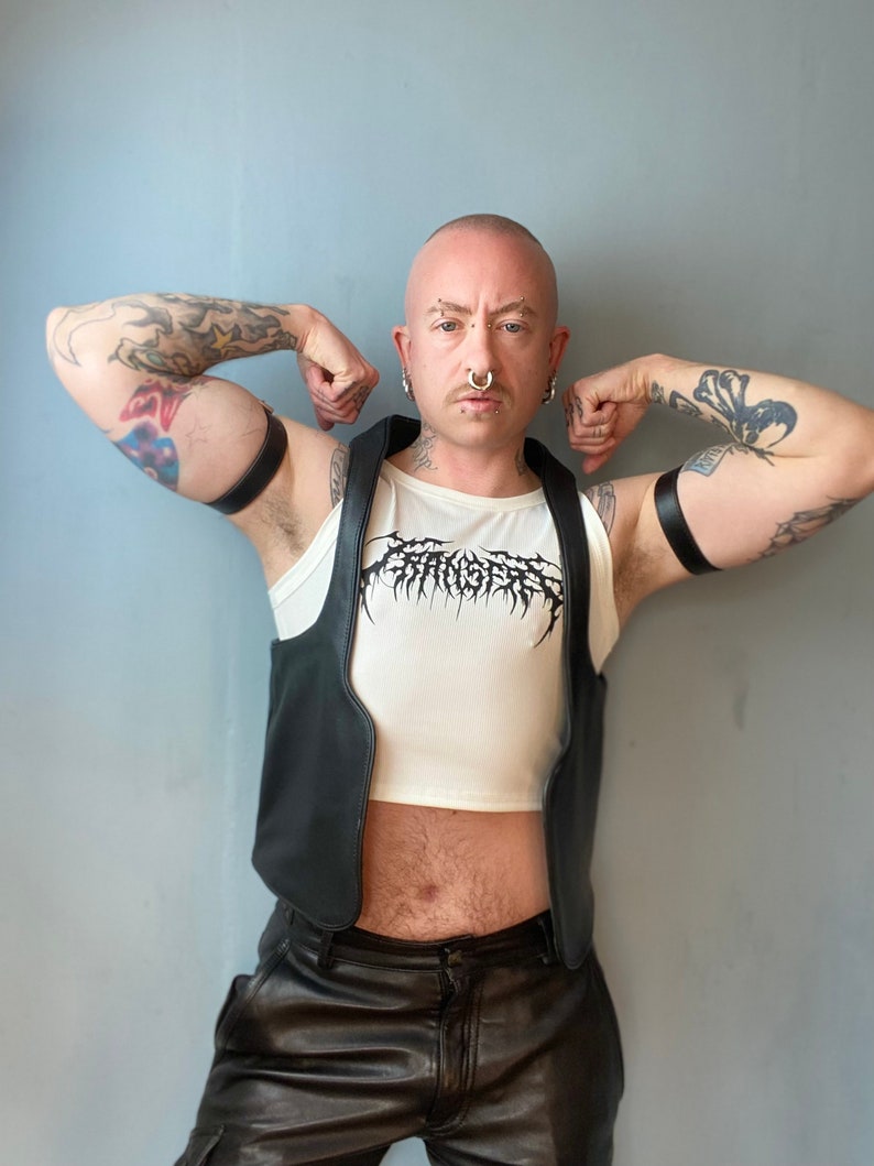 Op de afbeelding: Een persoon draagt een zwart leren vest over een wit crop top met een zwarte grafische print. Ze dragen een zwarte leren broek en hebben tatoeages op hun armen.