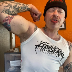 Op de afbeelding: Een wit tanktop met een zwarte grafische print van het woord "Cerberus" in een gotisch lettertype. De persoon die het tanktop draagt, draagt ook een zwarte beanie met het woord "Cerberus" in rode letters.