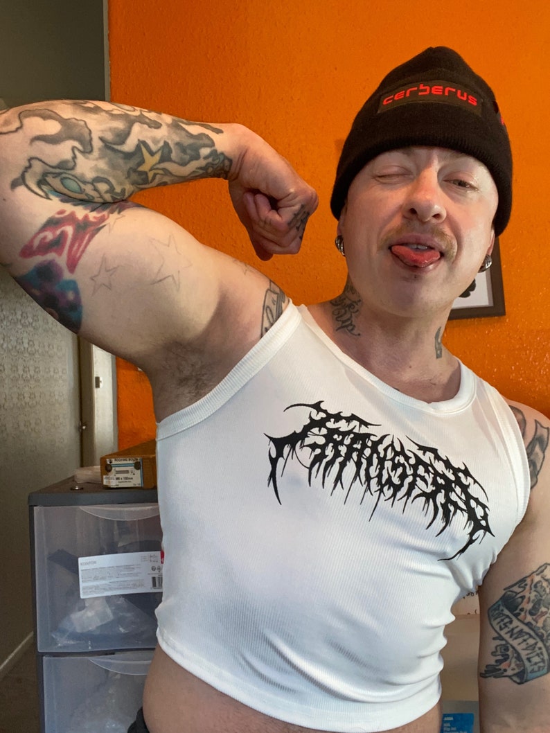 Op de afbeelding: Een wit tanktop met een zwarte grafische print van het woord "Cerberus" in een gotisch lettertype. De persoon die het tanktop draagt, draagt ook een zwarte beanie met het woord "Cerberus" in rode letters.
