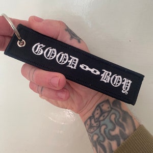 Puede incluir: Llavero de tela negro con texto bordado en blanco que dice "GOOD BOY" con un diseño de eslabón de cadena.