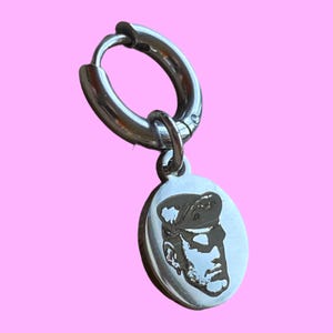May include: A silver hoop earring with a circular pendant. The pendant has a detailed engraved portrait of a person wearing a hat. The earring is set against a pink background.