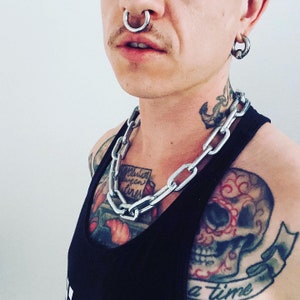 May include: A person with a silver nose ring and ear piercings is wearing a silver chain necklace with a large link design. The person has tattoos on their neck, chest, and arm. The tattoos include a skull, a heart, and the words "rebellion queen" and "a time".
