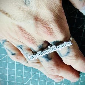 May include: A silver-toned metal ring featuring the text The Trans Agenda in a gothic-style font. The ring is designed to span across two fingers, with the lettering positioned horizontally along the top of the finger joints.