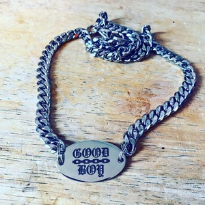 May include: A silver chain necklace with a silver oval pendant that says "GOOD BOY" in a gothic font. The pendant has a small chain detail.