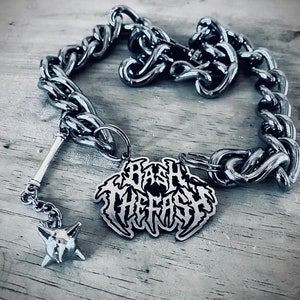 Könnte beinhalten: Eine silberne Kettenkette mit einem silbernen Anhänger, auf dem in schwarzen Buchstaben "Bash The Trash" steht. Der Anhänger ist an einem silbernen Dorn-Schlüsselanhänger befestigt.