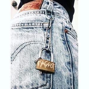 Op de afbeelding: Een zilveren ketting met een hangslot eraan. Het hangslot is van messing en heeft het woord "FAGGOT" in het zwart erop geschreven.