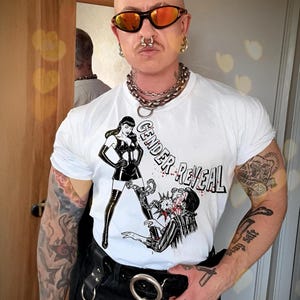 Peut inclure: Un t-shirt blanc avec une illustration graphique représentant une personne en tenue noire debout au-dessus d'une autre, avec le texte GENDER REVEAL imprimé dans une police stylisée. Le motif inclut des accents rouges et des détails à l'encre noire.