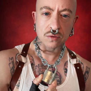 May include: A person wearing a white tank top, brown leather harness, and a silver chain necklace with a padlock. The person has multiple piercings and tattoos. The padlock has the text "5001 2001".