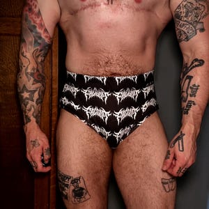 May include: Black high-waisted underwear featuring a repeating white graphic print of the word FAGGOT in a stylised, jagged, metal-inspired font. The garment has a fitted silhouette with a smooth fabric texture and a clean, finished waistband.