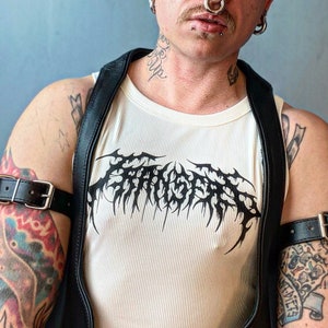 Op de afbeelding: Een wit tanktop met een zwarte grafische print van een gestileerd ontwerp. Het tanktop wordt gedragen door een persoon met tatoeages en leren banden op hun armen.