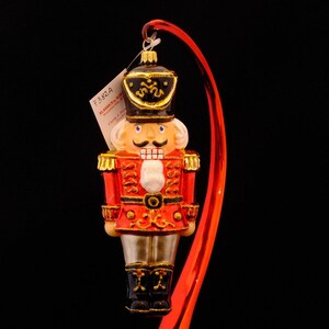May include: A red and gold glass Christmas ornament depicting a nutcracker soldier. The ornament has a black hat with gold trim and a white beard. The ornament is hanging from a red ribbon.