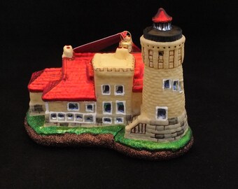 May include: A glass Christmas ornament shaped like a lighthouse with a red roof and a beige tower. The lighthouse has a small green lawn and a brown base.
