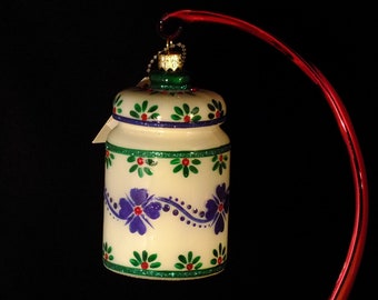 May include: A white glass Christmas ornament shaped like a jar with a blue and green floral design. The ornament has a red and green glitter trim.
