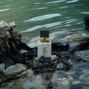 Può includere: Un flacone di profumo rettangolare in vetro con liquido ambrato e tappo nero, inserito in una base di cemento grigio chiaro. La base presenta un simbolo scuro inciso sulla parte anteriore. Il flacone è posizionato su una roccia scura.
